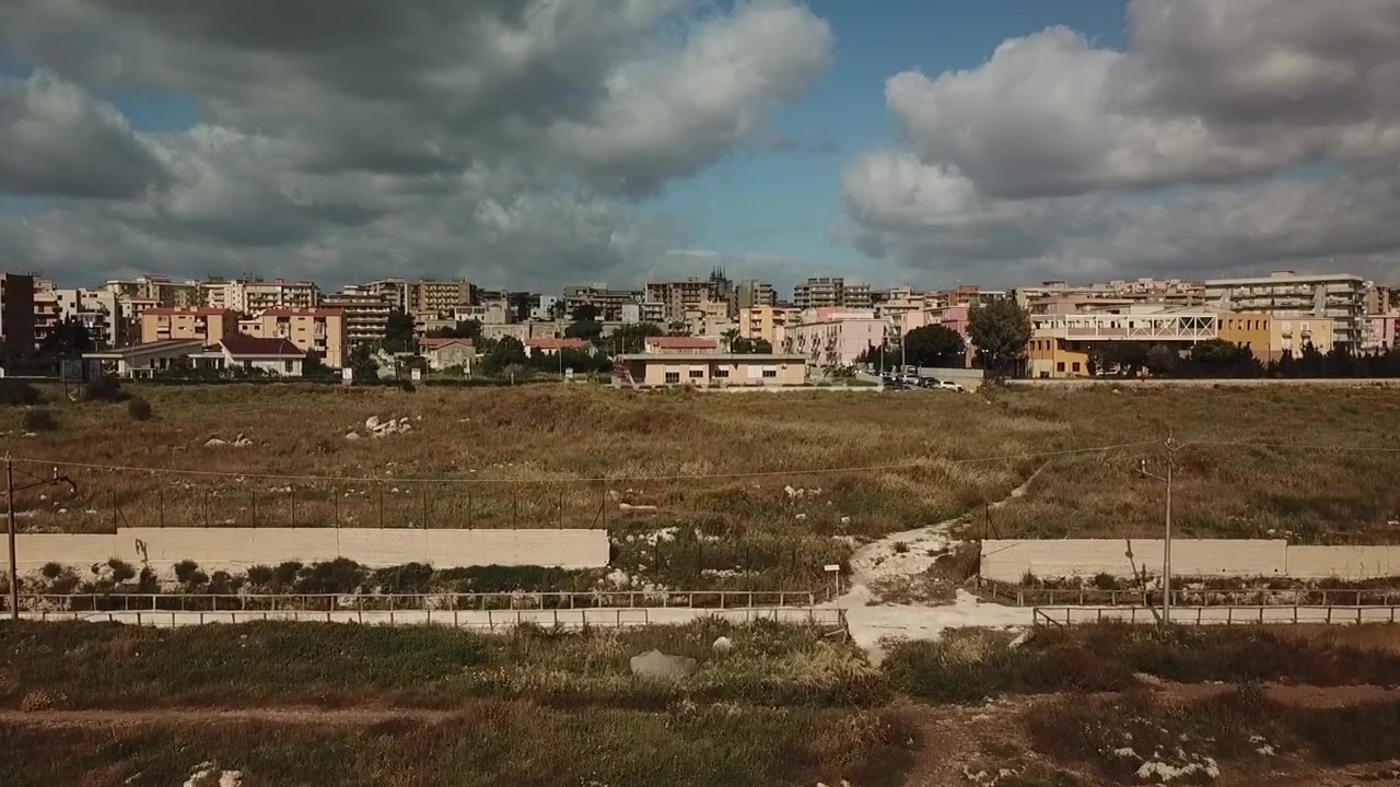 Tomada de drone em ascensão lenta revelando terrenos abertos, estradas e edifícios residenciais nos arredores de Siracusa, Sicília, sob nuvens dramáticas