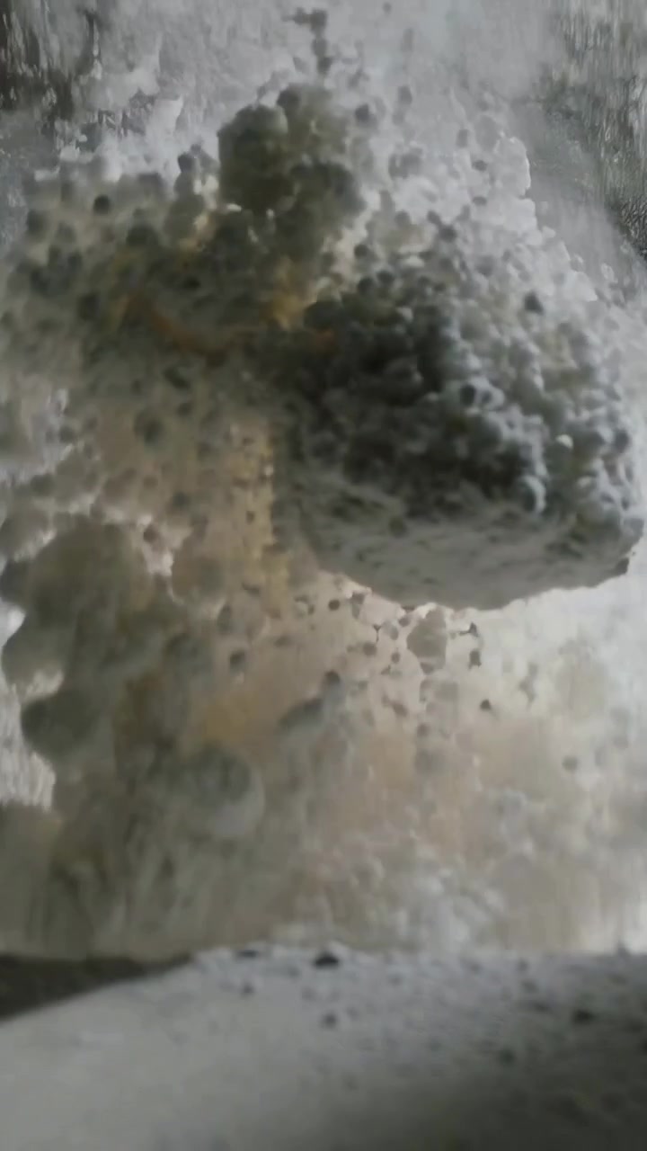 Vertical video: Releasing churning water flow down outlet lip, creating foam, spray and sediment