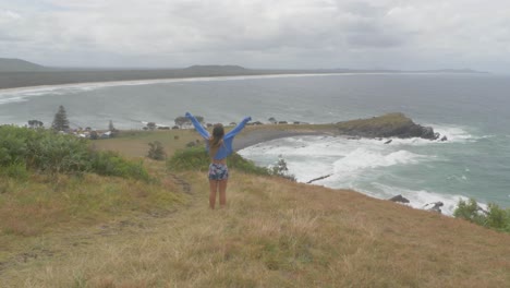 sexy girl raising arms in the air while feeling ocean breeze in the morning - ocean waves crashing at beach and peninsula at crescent head, nsw, australia