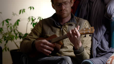 a guy playing a ukulele with a plant in the background and a light to his right