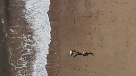 Drohnenaufnahme-Einer-Familie-Im-Urlaub,-Die-Am-Strand-Entlang-Läuft-Und-Wellen-Bricht