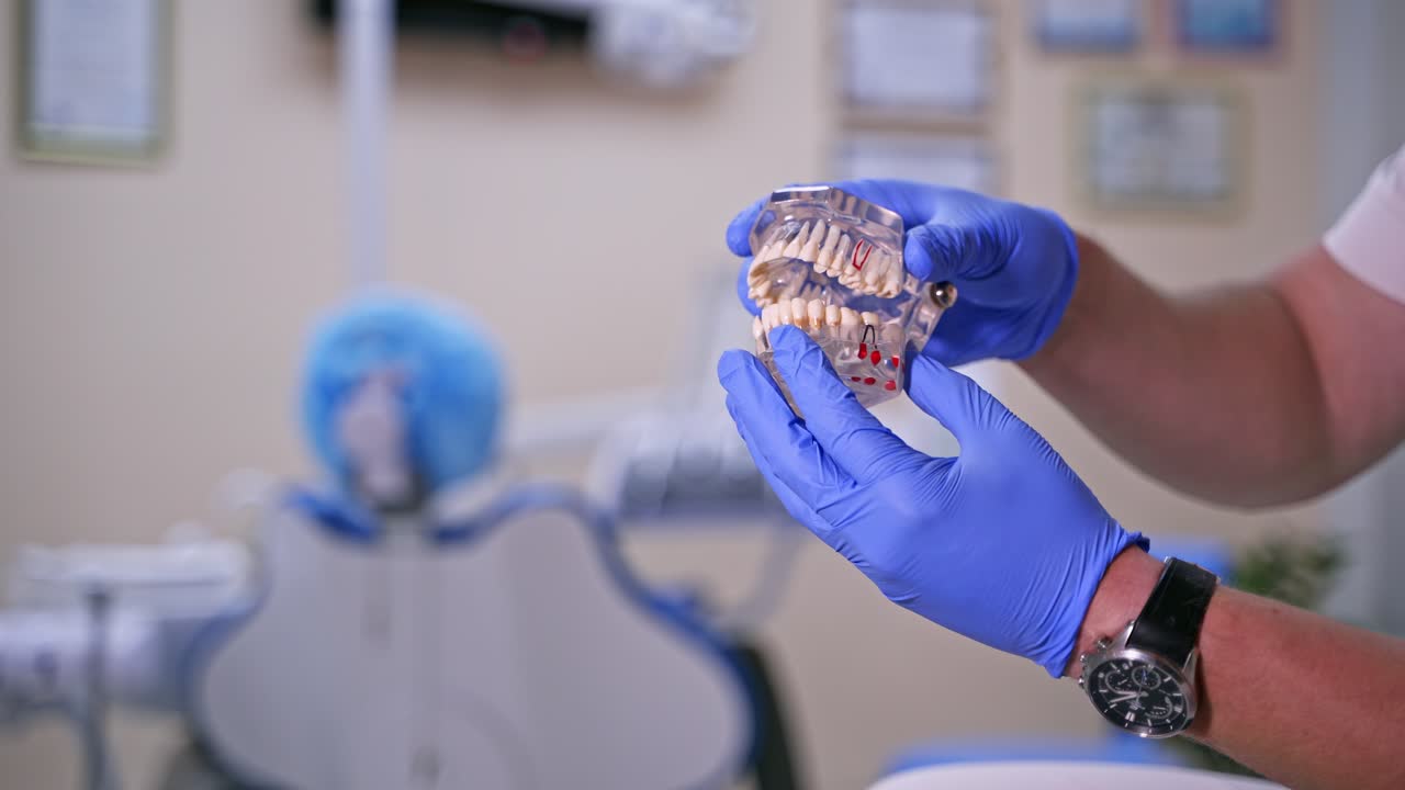 Doctor shows on a plastic jaw sample or model different methods of teeth treatment. Modern dental clinic background. Blue medical gloves on male doctor's hands. Dental Hygiene and Health concept video.