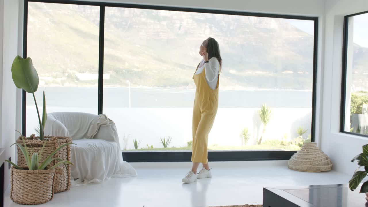 Escuchando una melodía suave, mujer baila frente a la ventana con movimientos de brazos celebrando la vista al lago.