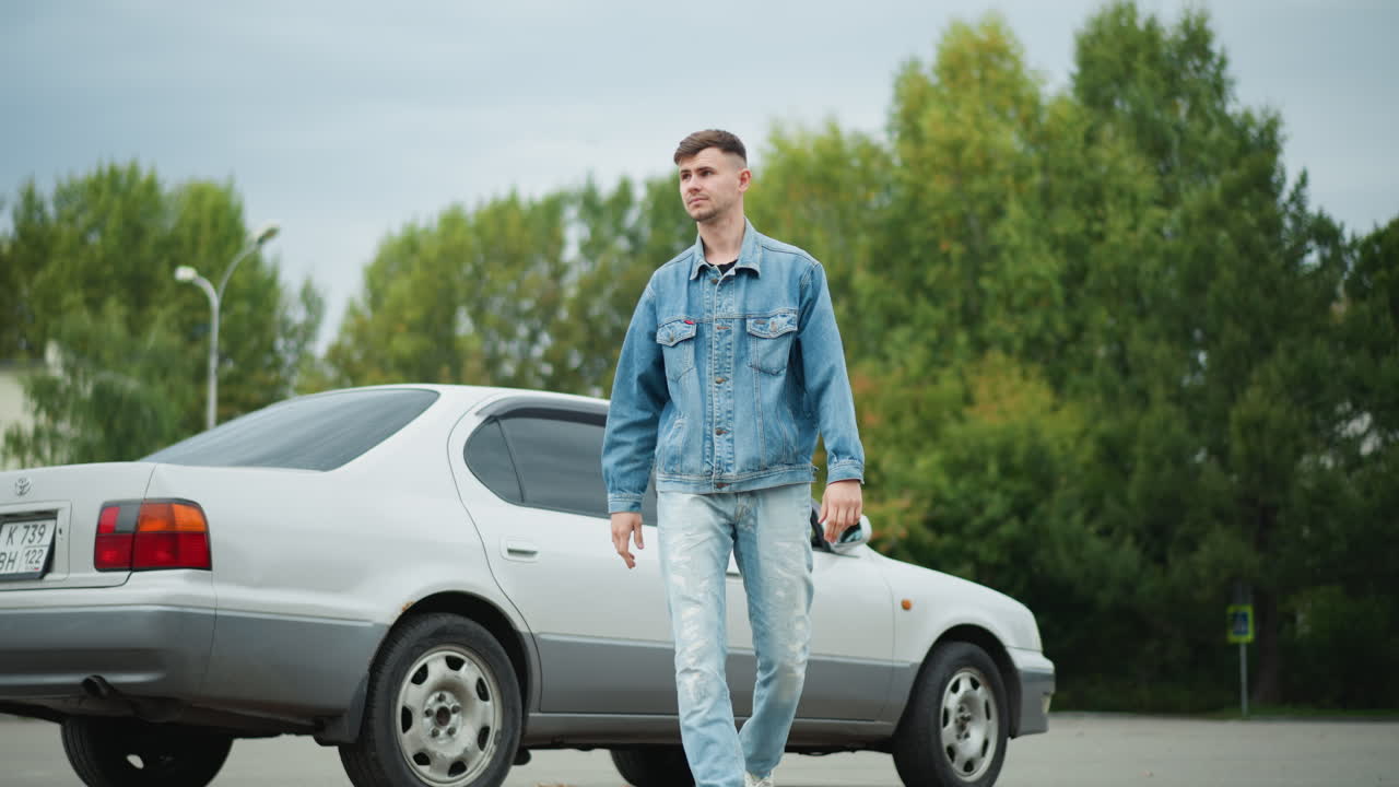 Giovane uomo bianco che scende da una berlina in un parcheggio suburbano vuoto, indossa una giacca di jeans e jeans strappati, cammina oltre gli alberi con passo sicuro, luce nuvolosa che crea un’atmosfera cinematografica