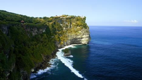 Slow-motion-reveal-shot-of-the-cliffs-of-Pura-Uluwatu-temple-in-Bali,-Indonesia-overlooking-the-beautiful-cliffs-and-high-waves-from-the-blue-sea