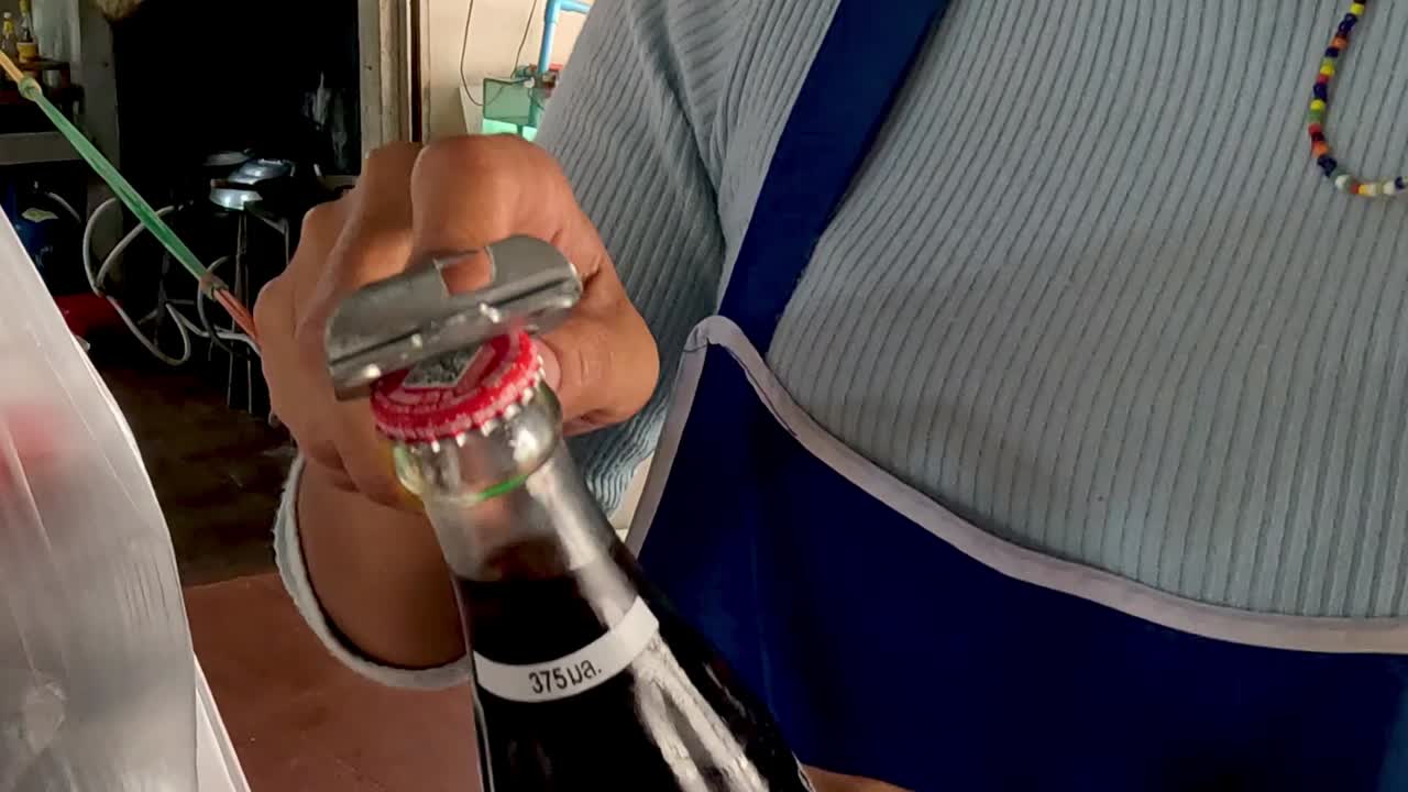 A person uses a metal bottle opener to remove the cap from a glass soda bottle in a casual setting.