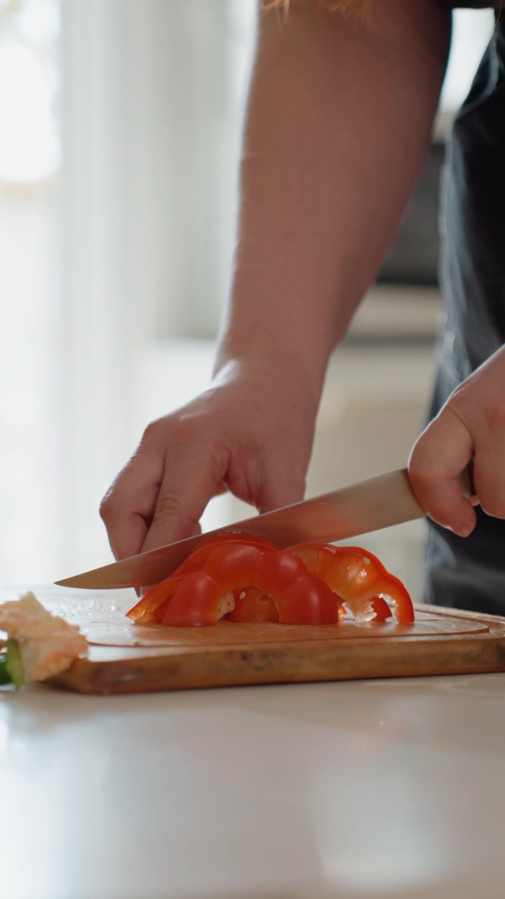 Primo piano di mani adulte che affettano un pomodoro maturo a fette sottili su un tagliere di legno con un coltello da chef; una mano tiene fermo il frutto mentre l'altra preme la lama verso il basso, mostrando un'attenta preparazione in cucina.