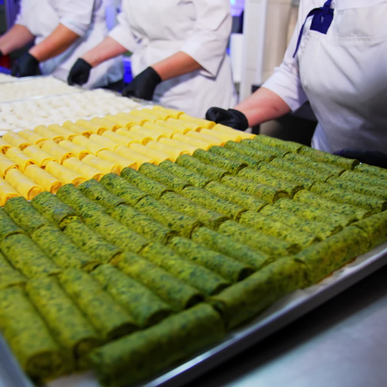 Three cooks put the big trays with semi-cooked food on the metal table. Stuffed pancakes and meat ravioli manufactured at food factory