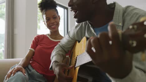 video of happy african american couple sitting on sofa, singing and playing guitar