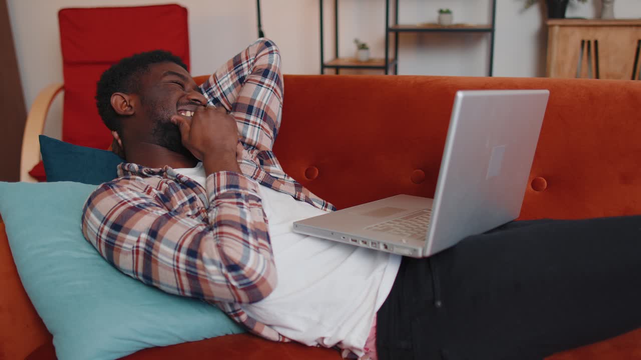 Young man using laptop computer lying on sofa at home working online shopping watching movies