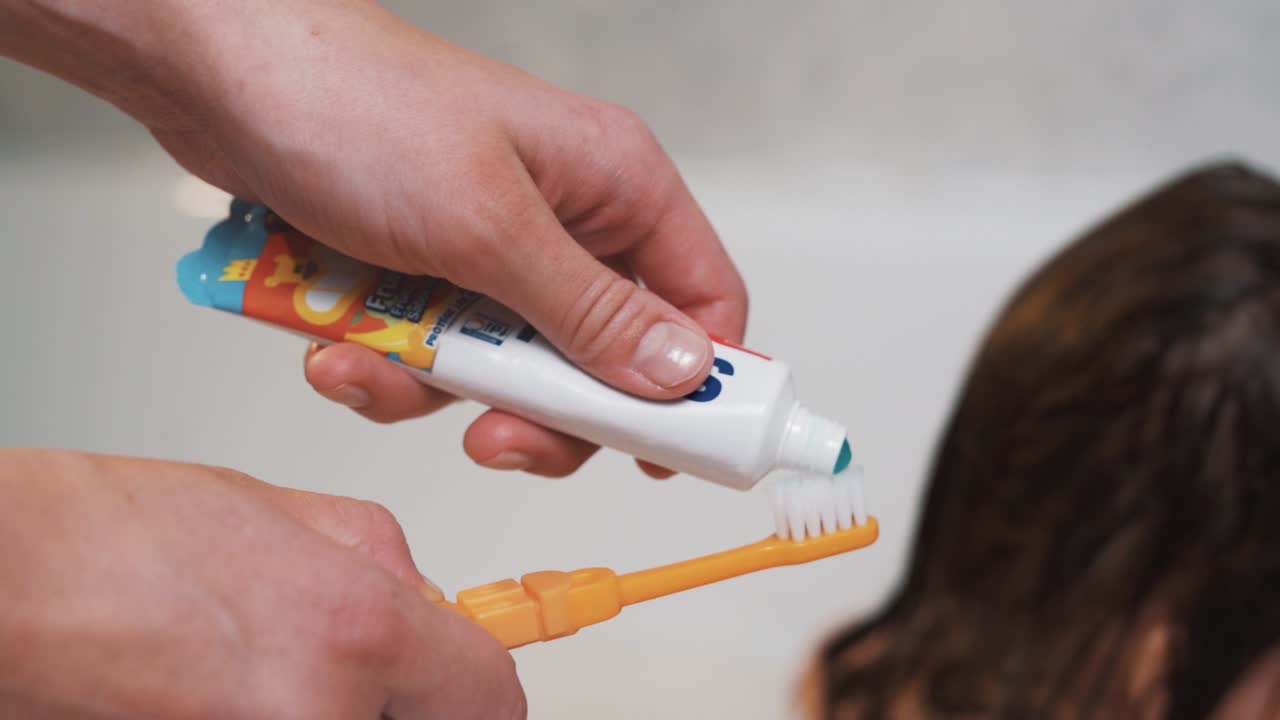 Close-up of female hands holding a child's toothbrush and applying toothpaste on the background of a child in a bath