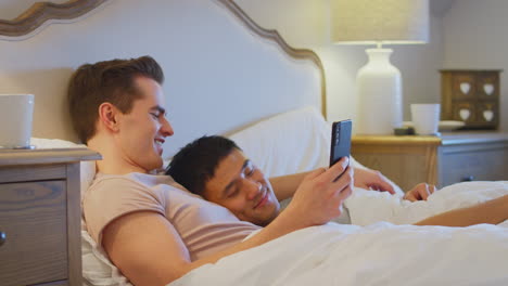 loving same sex male couple lying in bed at home looking at social media on mobile phone