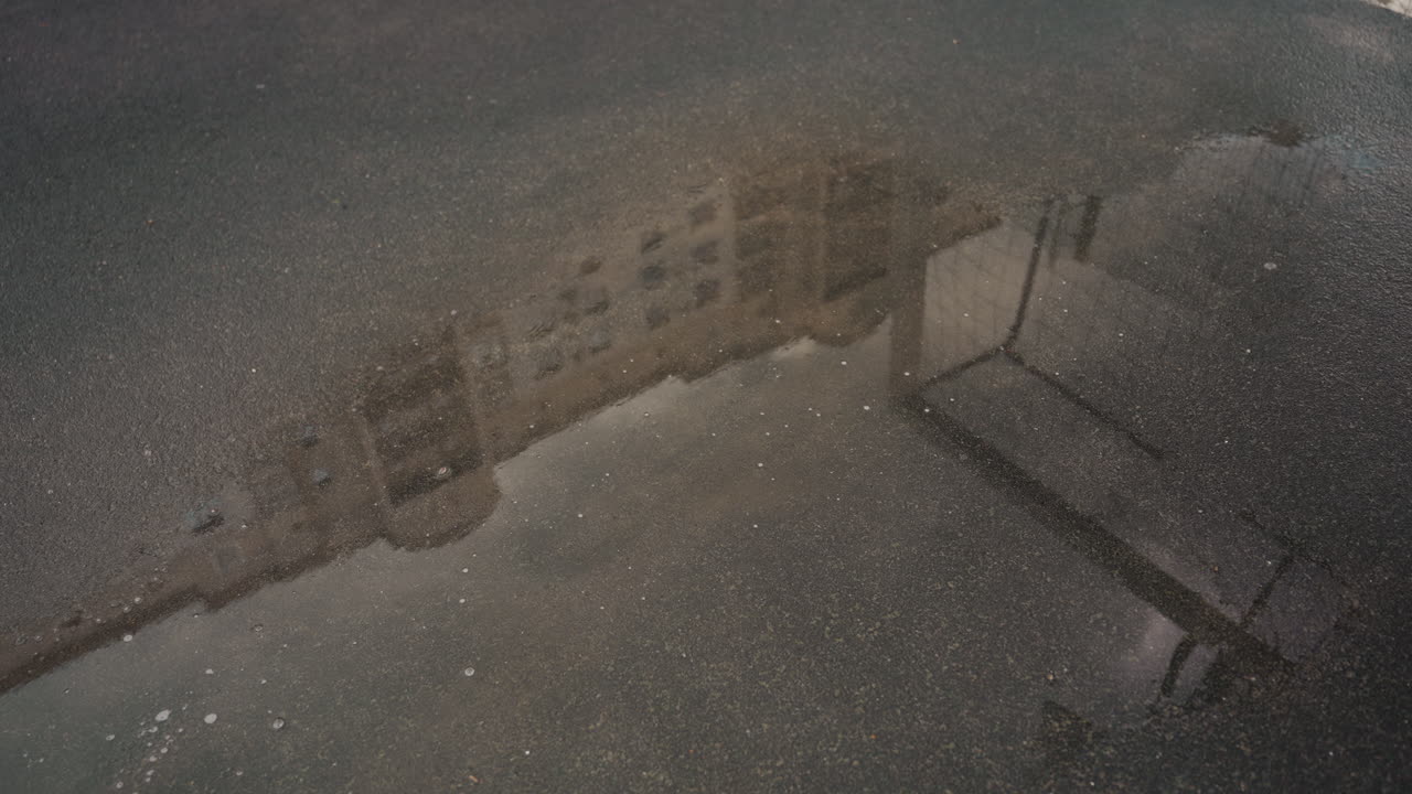 Puddle reflection of street and ball, moody urban scene after rain, soccer ball near sneaker, foot midstep creating small splash, building reflection on wet asphalt, overcast evening light, candid