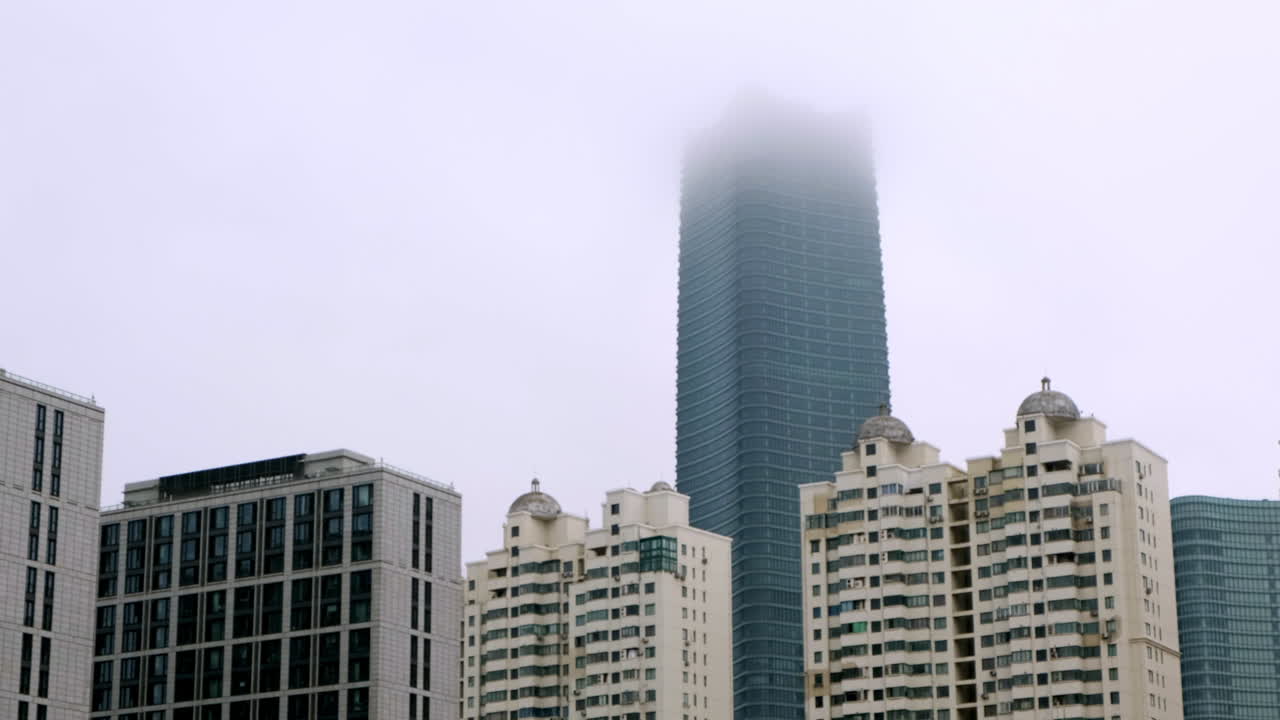 Um imponente arranha-céu moderno de vidro desaparece em nevoeiro branco denso acima de um aglomerado de edifícios residenciais altos em Xangai