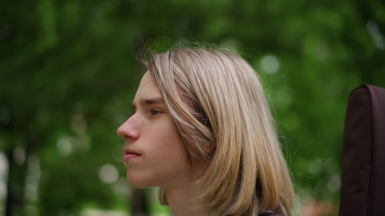 giovane chitarrista bianco in un parco che trasporta una custodia per chitarra sulla spalla, pensieroso musicista adolescente con capelli biondi, ritratto spontaneo tra alberi verdi, luce pomeridiana soffusa, abbigliamento casual, stato d'animo riflessivo