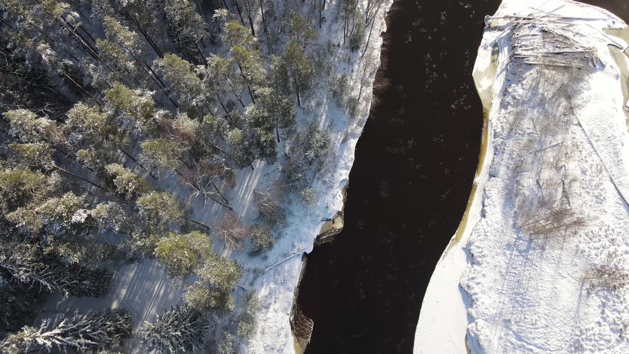 Aerial view of a winding river through a dense snow-covered forest in winter, with a curving road running alongside. A serene and scenic natural landscape.