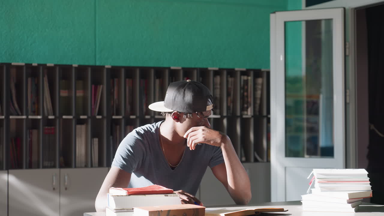 Close up of man in casual t-shirt and backward cap sitting indoors with hand resting on chin, gazing thoughtfully to side near stack of books in bright sunlight with blurred bookshelf in background