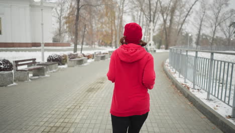 back view of woman jogging along snowy park pathway in red hoodie, surrounded by serene winter scenery with benches, bushes, iron railing, lamp posts, white building, and distant cross monument