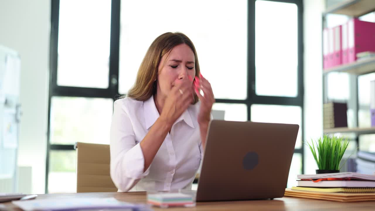 Woman working in office, sneezes, and rubs her eyes