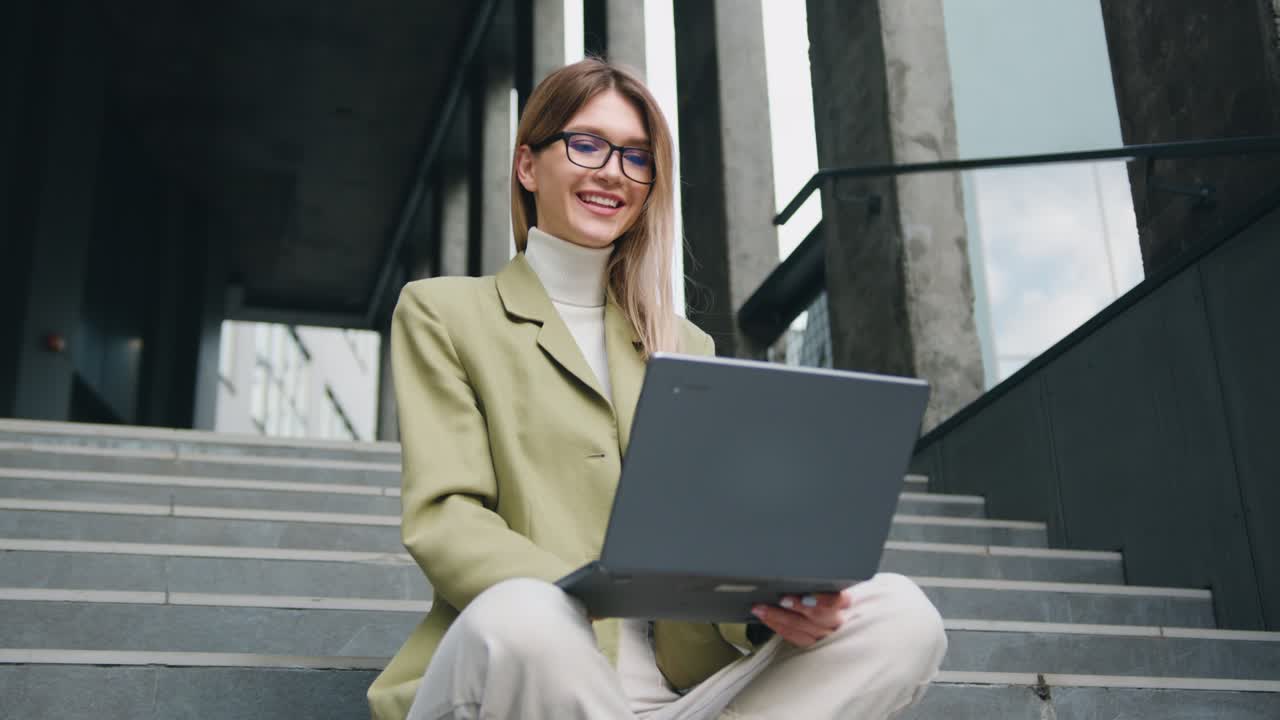 Good -looking young caucasian woman in glasses wearing a green jacket sitting with crossing legs on the stairs outside, typing on her laptop with satisfied face
