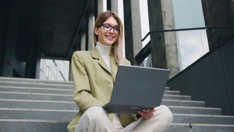 good -looking young caucasian woman in glasses wearing a green jacket sitting with crossing legs on the stairs outside, typing on her laptop with satisfied face
