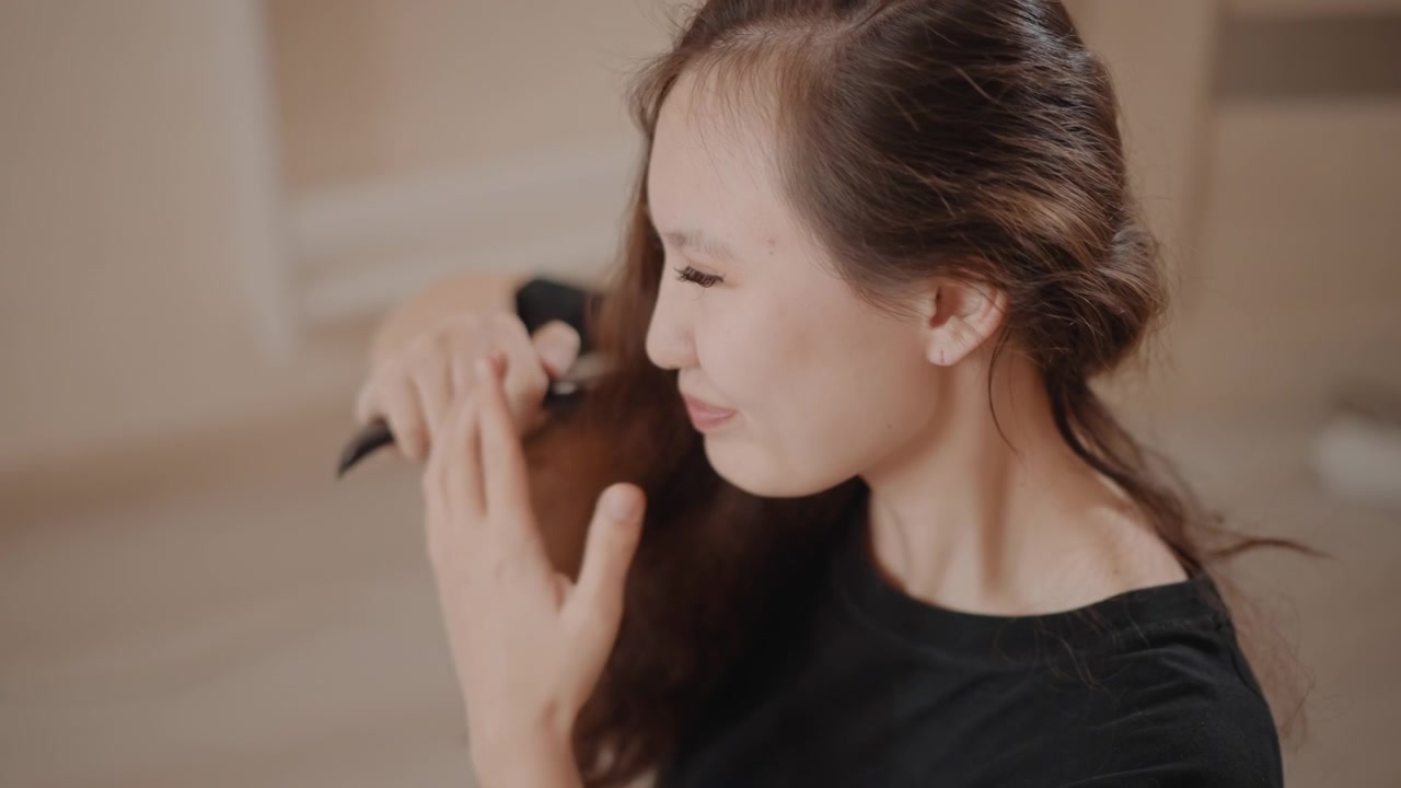 若い女性が室内でヘアケア中に座り、ブラシを使って長い黒髪の絡まりを一定の動きで解いている。髪の根元から毛先まで、その動作が見て取れ、なめらかで艶やかな仕上がりになっている。