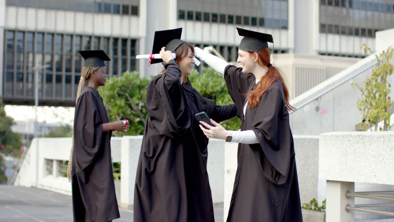 Donna dai capelli rossi che alza lo smartphone per avviare una chiamata di gruppo con amici che tengono diplomi su un vialetto