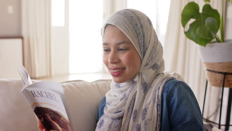 Happy-biracial-woman-in-hijab-reading-book-on-sofa-at-home-with-copy-space,-slow-motion