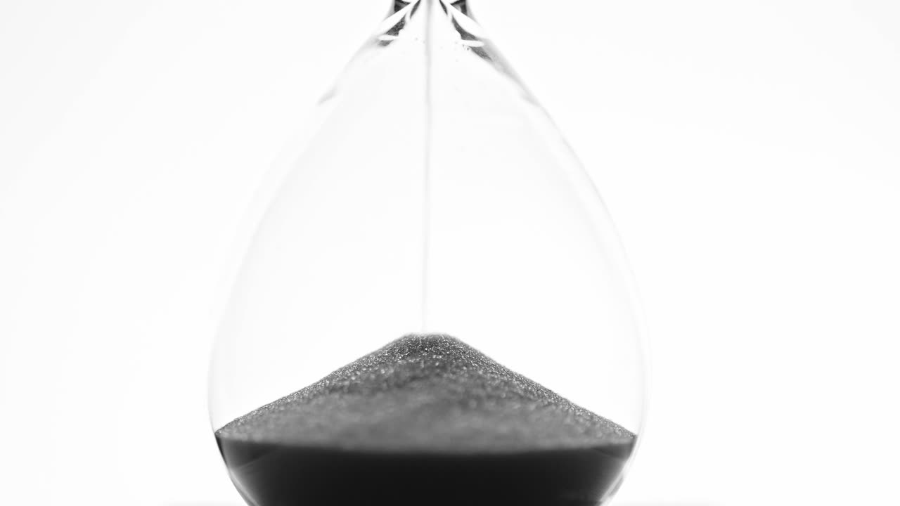 A close-up of a minimalist hourglass with black sand flowing down, symbolizing the passage of time. The high-key lighting and white background emphasize the simplicity and elegance of the design.