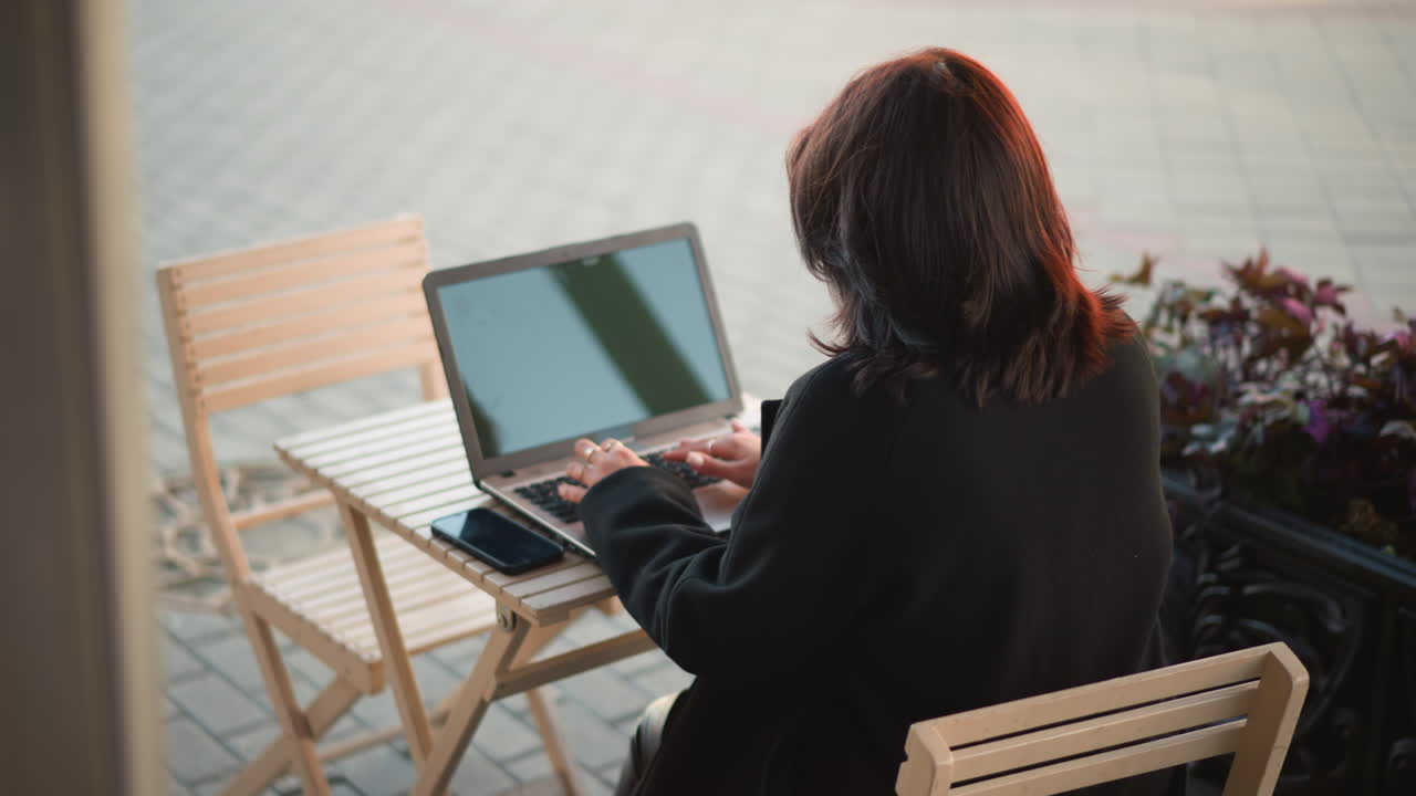 Freelancer seated on wooden chair typing on laptop with smartphone nearby on table, outdoor setting surrounded by plants, creating a peaceful and focused work environment