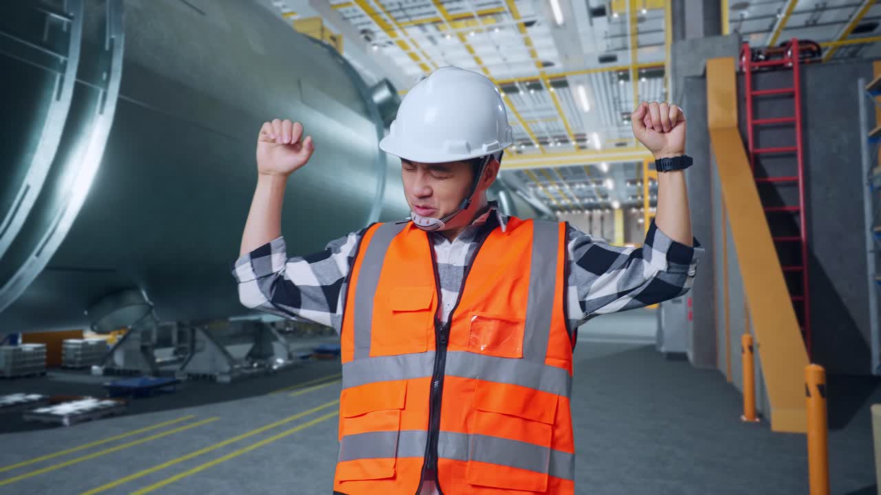 안전 헬을 입은 아시아 남성 엔지니어가 파이프 제조 공장에서 서서 춤을 추며 축하하고 있습니다.