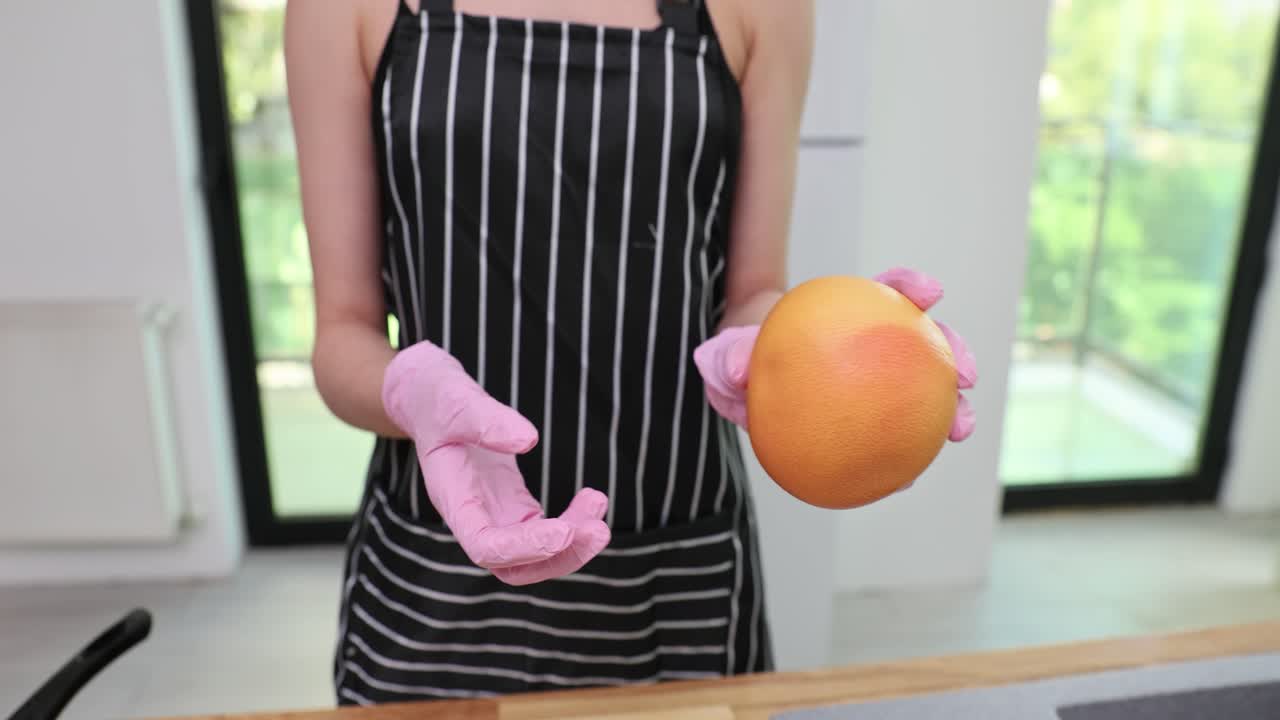 Person in apron and pink gloves holding a grapefruit in a kitchen