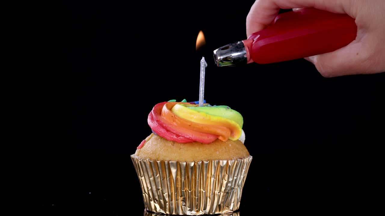 Una mano usa un accendino rosso per accendere una singola candela sopra un cupcake glassato arcobaleno, in un ambiente scuro illuminato da studio con uno sfondo nero.
