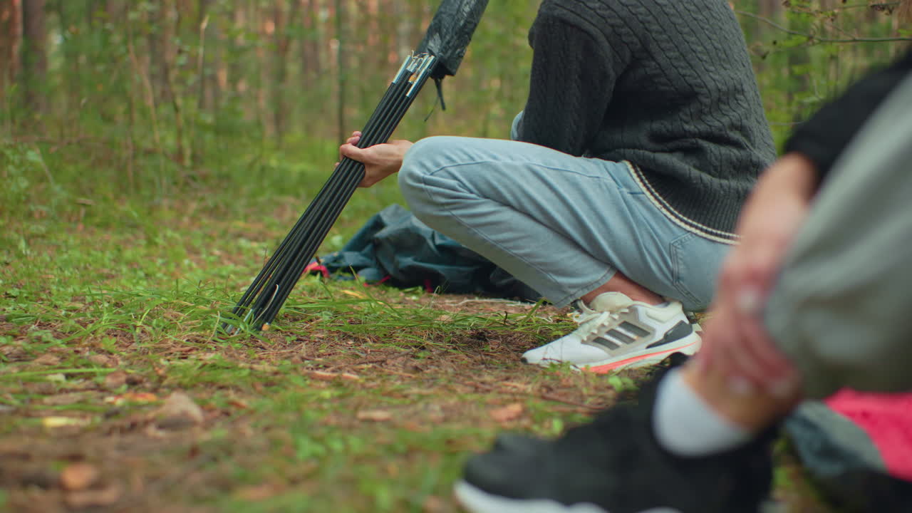 Close up of boy squatting on forest ground carefully pulling tent pole from bag while person in foreground watches attentively during camping setup in green wooded outdoor environment