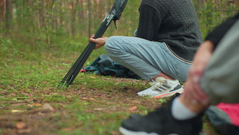 close up of boy squatting on forest ground carefully pulling tent pole from bag while person in foreground watches attentively during camping setup in green wooded outdoor environment