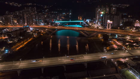 Drohnen-Nacht-Hyperlapse-Einer-Asiatischen-Stadt-Mit-Gebäuden,-Brücke,-Fluss,-Autos-Und-Bunten-Lichtern