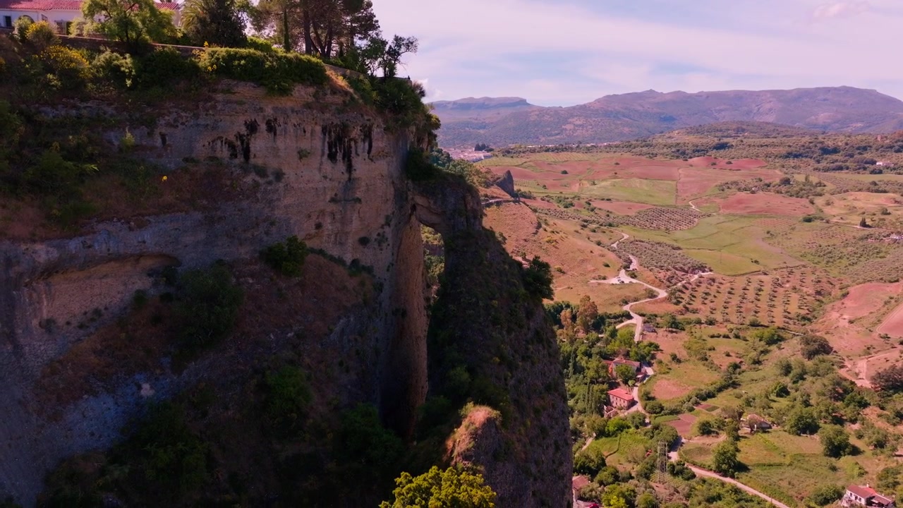Imágenes de dron descendiendo junto a la imponente pared del acantilado de Ronda, revelando un arco natural tallado en la pared rocosa y un extenso valle bañado por el sol de olivares y carreteras sinuosas abajo.