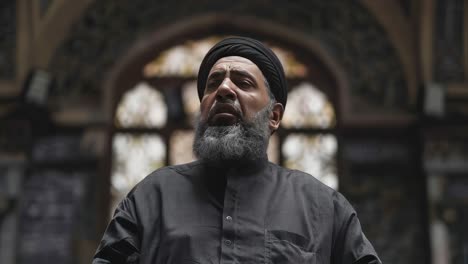 Bearded religious leader wearing black clothes and a kufi, looking up and reciting islamic prayers inside a mosque with stained glass windows