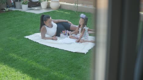 a mother and her young daughter sit on a blanket in the backyard, engaging in a drawing activity together. the outdoor scene captures a peaceful family moment, fostering creativity and bonding.