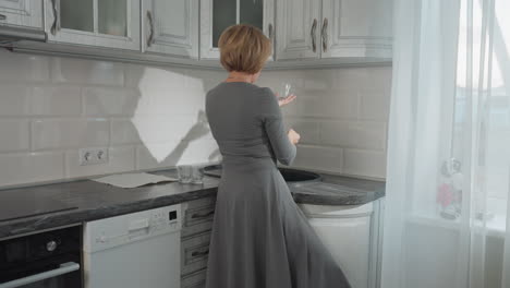 back view of lady in plaid gown holding glass cup while dancing happily in bright kitchen with two other glass cups on marble countertop, sunlight streaming through white curtain nearby window