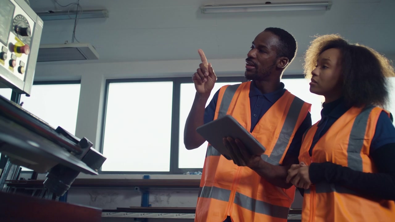 Lavoratori in un impianto industriale, un dipendente senior che tiene in mano un tablet digitale e spiega a un nuovo collega.