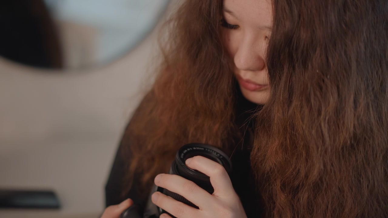 Vista cercana de una persona con cabello largo y castaño manipulando cuidadosamente un objetivo de cámara, girando el barril de la lente con ambas manos mientras mira hacia abajo con atención concentrada, junto a equipo de mesa y suave.