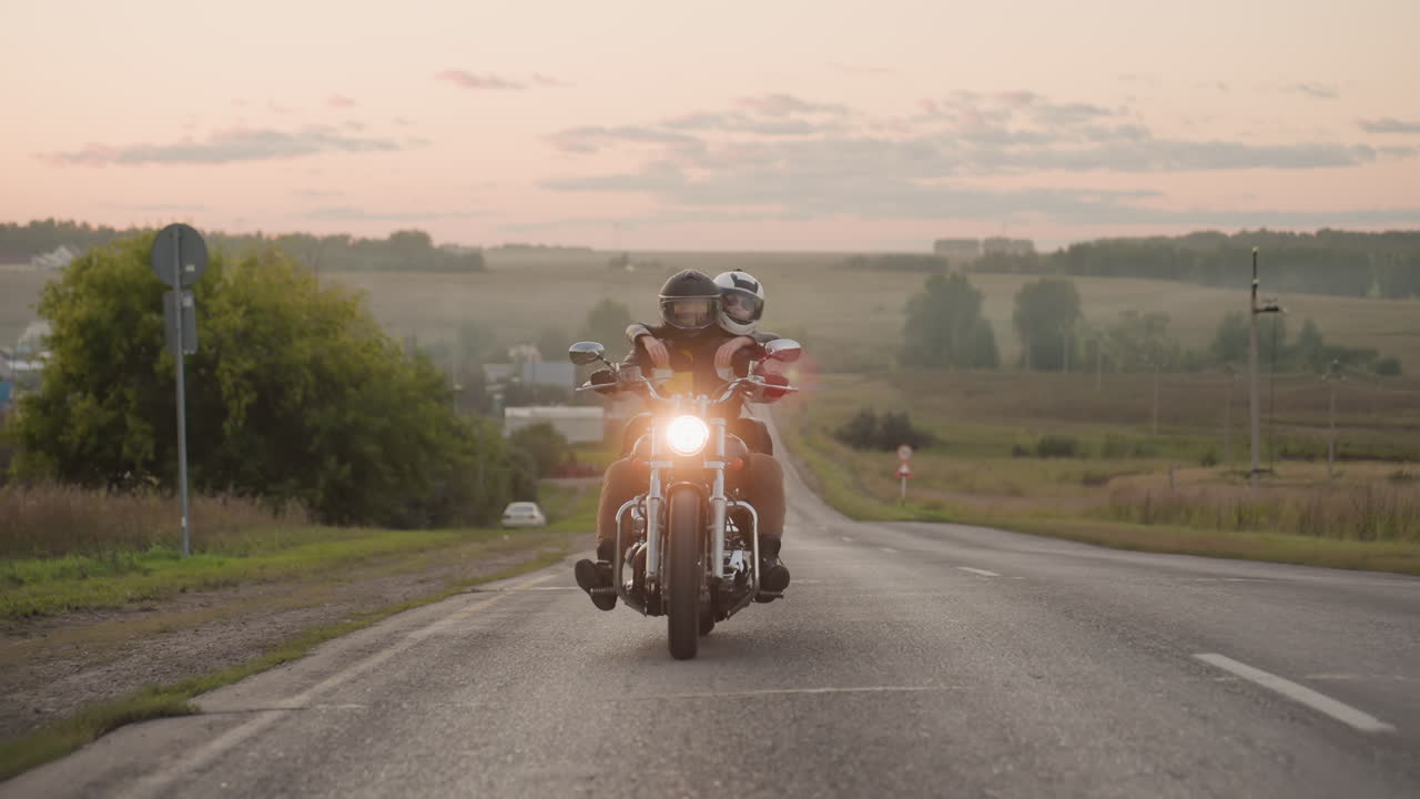 Motorcyclist with passenger rides uphill on countryside road during golden sunset, glowing headlight shining against scenic rural background with open fields, trees, horizon, and peaceful evening sky