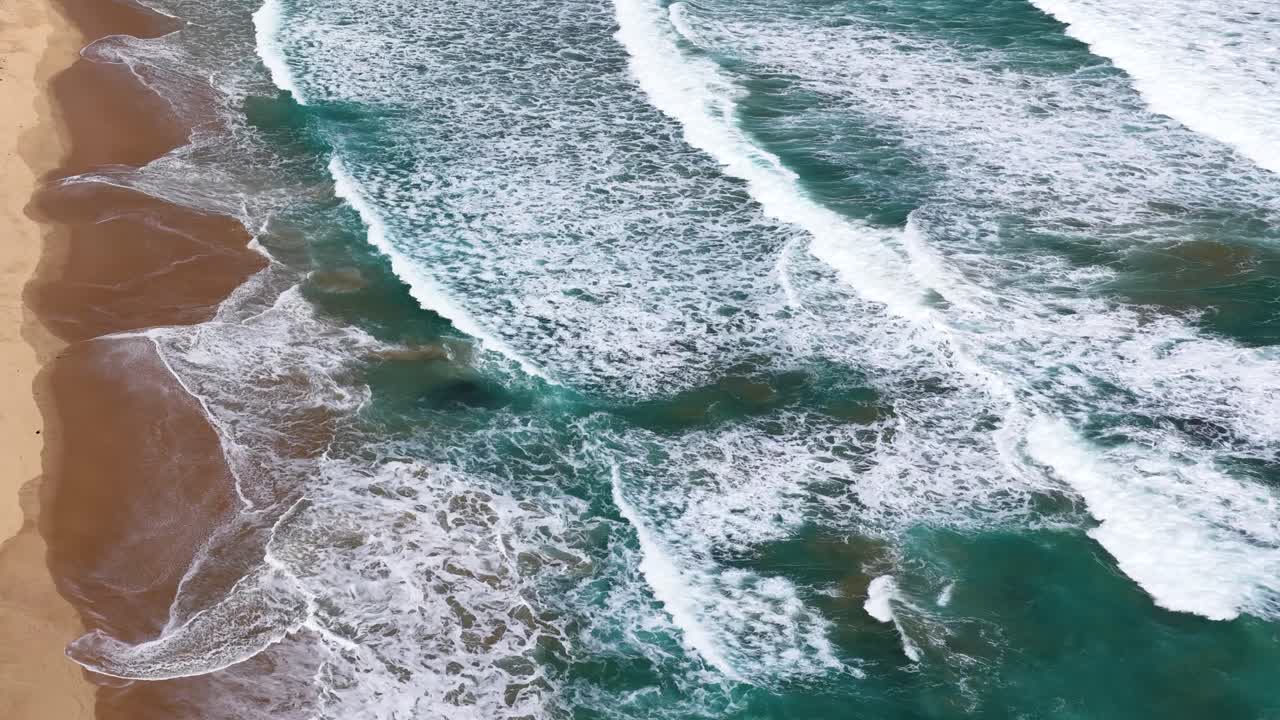 Drone captures turquoise waves breaking on sandy shore, natural daylight, smooth lateral camera movement