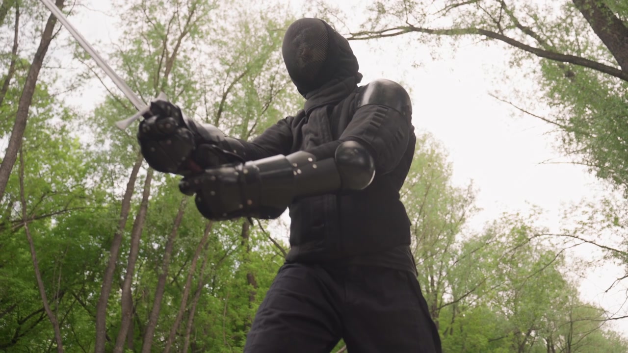 Close up of person in black fencing gear holding two swords demonstrating various stances and movements outdoors amidst green trees. subject face is covered by mesh protection while they