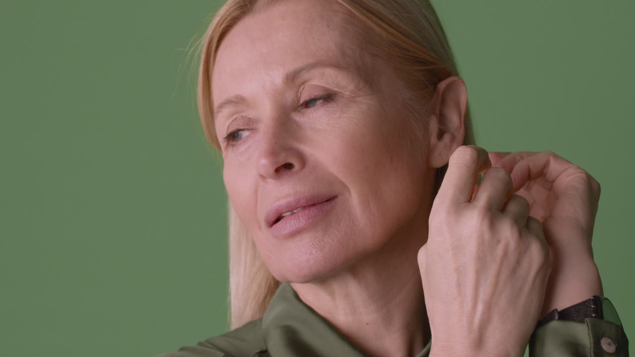 녹색 셔츠를 입은 금발의 성숙한 여인의 모습을 가까이서 보고, 옆을 보고 녹색 배경에 웃고 있는 그녀의 귀 뒤에 머리를 따는 것