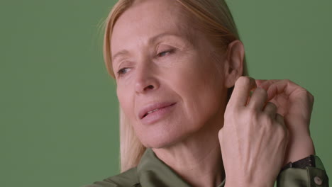 Close-Up-View-Of-Blonde-Mature-Woman-Wearing-Green-Shirt,-Picking-Her-Hair-Behind-Her-Ear-Looking-At-Side-And-Smiling-On-Green-Background