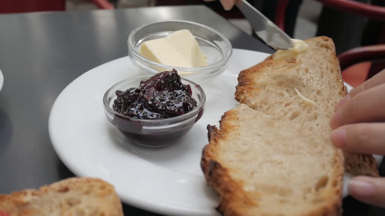Spreading butter on toast with jam in cozy outdoor café setting, close-up view
