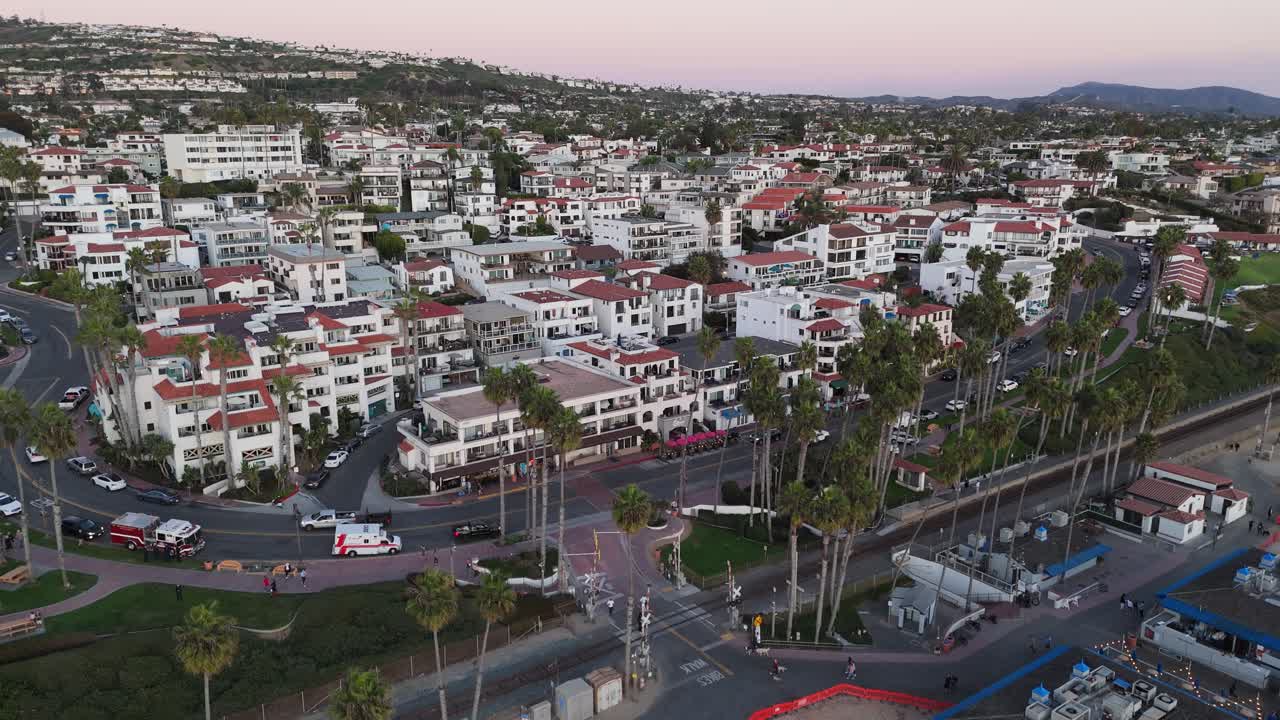 A dynamic drone push-in shot of the San Clemente Pier Bowl neighborhood, highlighting Spanish-style architecture, palm trees, and proximity to the beach. Captured at twilight.