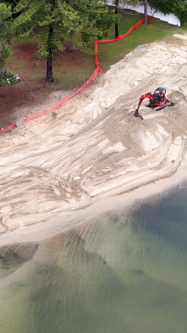 Inquadratura aerea dall'alto di macchinari pesanti che spostano sabbia per il ripascimento della spiaggia lungo un canale.